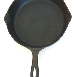May include: A black cast iron skillet with a long handle and a loop for hanging. The skillet has a round cooking surface and two small spouts on either side. The pan is made of heavy-duty metal.