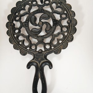May include: Black cast iron trivet with an ornate, openwork design. The circular trivet features a central swirl pattern and a decorative handle. The trivet is designed to protect surfaces from hot cookware.