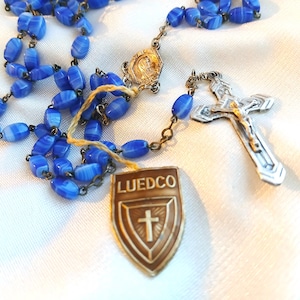 May include: A vintage rosary with blue oval beads and a silver crucifix. The rosary includes a shield-shaped pendant with the text "LUEDCO" and a cross. The beads are strung on a metal chain.