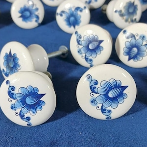 May include: White ceramic cabinet knobs with a blue floral design. The knobs are round with a glossy finish and a metal screw for installation. The blue floral pattern is hand-painted and features a stylized flower with delicate details.