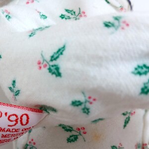 Pu&ograve; includere: Primo piano di un tessuto bianco con un motivo ripetuto di foglie di agrifoglio verdi e bacche rosse. Un'etichetta con il testo "'90" e "Made on" &egrave; visibile. Il tessuto &egrave; morbido, forse di un articolo natalizio.