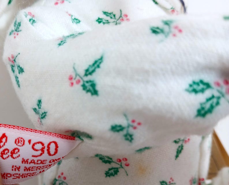 Pu&ograve; includere: Primo piano di un tessuto bianco con un motivo ripetuto di foglie di agrifoglio verdi e bacche rosse. Un'etichetta con il testo "'90" e "Made on" &egrave; visibile. Il tessuto &egrave; morbido, forse di un articolo natalizio.
