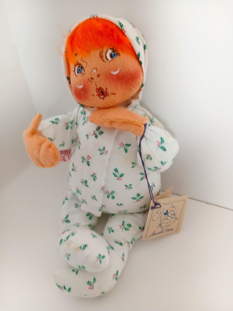Pu&ograve; includere: Una bambola vintage fatta a mano con capelli arancioni, occhi blu e guance rosee. La bambola indossa una tutina bianca con un motivo floreale verde e rosa e un cappuccio abbinato. Un'etichetta &egrave; attaccata alla bambola.