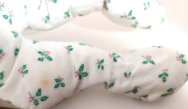 Pu&ograve; includere: Primo piano di un oggetto in tessuto bianco con un motivo di foglie di agrifoglio verdi e bacche rosse. Il tessuto sembra morbido e felpato. Ci sono alcune piccole macchie marroni chiare sul tessuto.