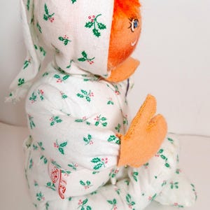 Pu&ograve; includere: Bambola vintage con un pigiama e un cappuccio bianchi, entrambi decorati con agrifoglio verde e bacche rosse. La bambola ha capelli arancioni, un viso arancione e mani arancioni. La bambola &egrave; seduta.