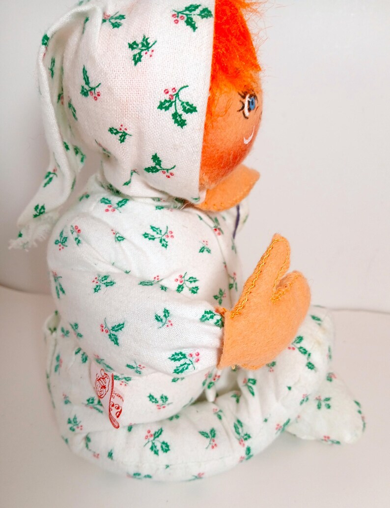 Pu&ograve; includere: Bambola vintage con un pigiama e un cappuccio bianchi, entrambi decorati con agrifoglio verde e bacche rosse. La bambola ha capelli arancioni, un viso arancione e mani arancioni. La bambola &egrave; seduta.