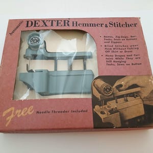 May include: A vintage Dexter Hemmer & Stitcher sewing machine in its original box. The box is a reddish-brown color with the words "DEXTER Hemmer & Stitcher" printed on the top. The machine is a light blue-grey color and is visible through a clear window.