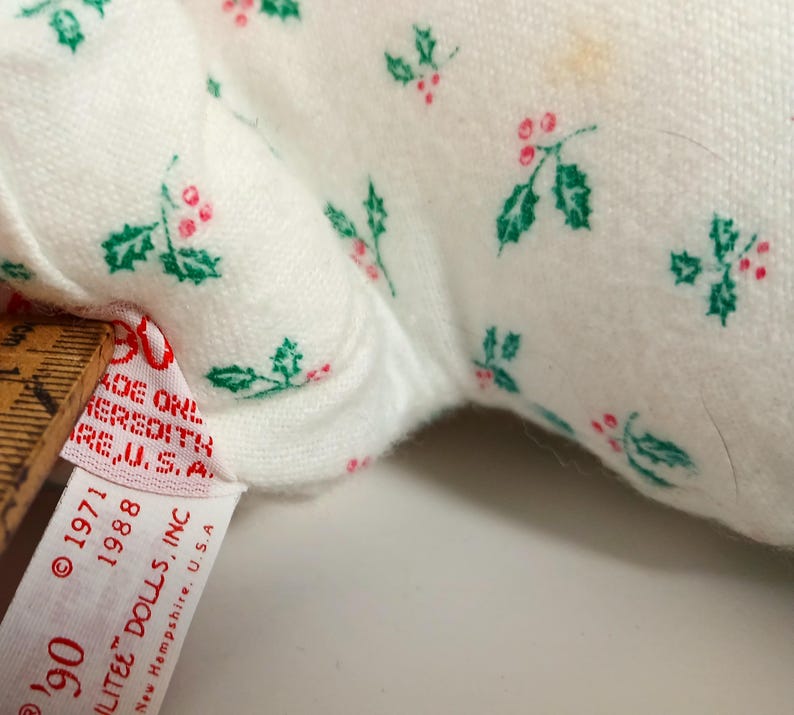 Pu&ograve; includere: Primo piano di un tessuto bianco con un motivo ripetuto di foglie di agrifoglio verdi e bacche rosse. Un'etichetta con testo tra cui "ILITEE DOLLS, INC" e "New Hampshire, U.S.A" &egrave; visibile. L'etichetta mostra anche le date di copyright del 1971 e 1988.