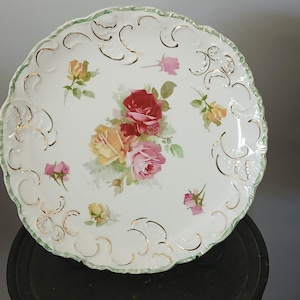 May include: A white porcelain plate with a scalloped edge and a green trim. The plate features a floral design with red, pink, and yellow roses. Gold accents are present throughout the design.