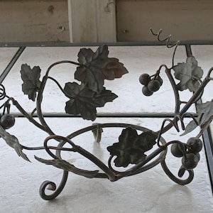 May include: A decorative metal fruit bowl with a grape vine design. The bowl features dark metal leaves, grapes, and curling vines. The bowl is supported by three small feet and is designed to hold fruit or other decorative items.