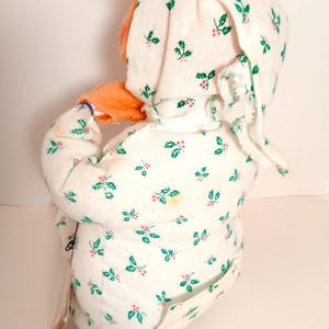 Pu&ograve; includere: Una bambola vintage che indossa una tutina bianca e un cappuccio abbinato, entrambi decorati con un motivo di agrifoglio verde e bacche rosse. La bambola ha i capelli arancioni e il viso arancione. La bambola &egrave; girata di spalle alla fotocamera.