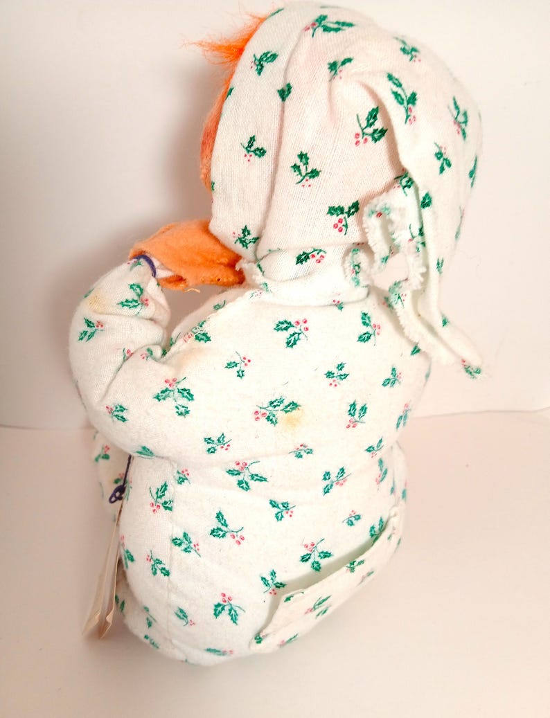 Pu&ograve; includere: Una bambola vintage che indossa una tutina bianca e un cappuccio abbinato, entrambi decorati con un motivo di agrifoglio verde e bacche rosse. La bambola ha i capelli arancioni e il viso arancione. La bambola &egrave; girata di spalle alla fotocamera.