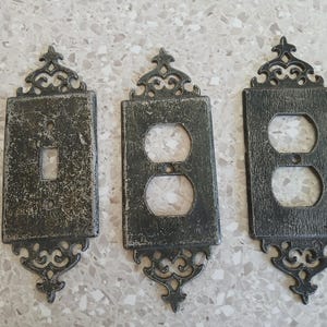 May include: Three rectangular wall plates with a dark, distressed metallic finish and ornate, scrollwork detailing at the top and bottom. One plate features a single toggle switch opening, while the other two plates feature dual outlet openings.