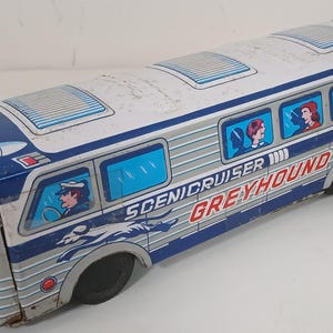 May include: Vintage tin toy bus in silver, blue, and red. The bus features the words "Scenicruiser Greyhound" and illustrations of passengers and a driver. The toy has black wheels and is likely from the mid-20th century.