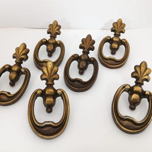 May include: A set of six brass-toned metal drawer pulls, each featuring a decorative fleur-de-lis top ornament and a swinging oval ring handle. The hardware has a warm, aged metallic finish with visible highlights and shadows on the surfaces.