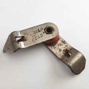 May include: Vintage metal can opener with the text "Quad Fold" stamped on the surface. The can opener is silver with rust spots and a red rubber wheel. The can opener is open and angled.