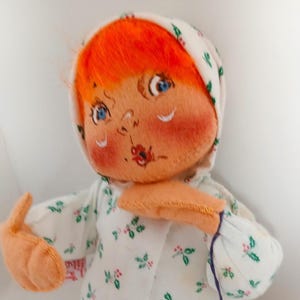Pu&ograve; includere: Bambola fatta a mano con capelli arancioni, occhi blu e guance rosee. La bambola indossa un cappuccio bianco e un vestito bianco con un motivo floreale verde e rosso. Le braccia della bambola sono tese e il viso &egrave; dipinto a mano.