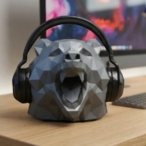 Könnte beinhalten: Ein grauer, geometrischer Bären-Kopfhörerständer mit schwarzen Kopfhörern. Der Ständer besteht aus eckigen Formen und hat einen offenen Mund. Die Kopfhörer sind schwarz und haben ein gepolstertes Kopfband.