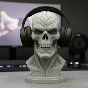 May include: A gray skull-shaped headphone stand with a pair of black headphones. The stand has detailed facial features and sits on a round base. The background includes a computer monitor, keyboard, and speaker.