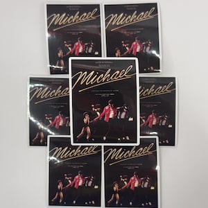 May include: A collection of rectangular stickers featuring the name "Michael" in gold script. The stickers showcase a group of people in a performance, with a central image of a person in a red jacket. The background is dark.