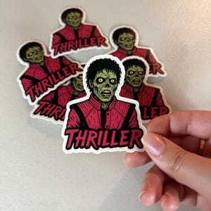 Może przedstawiać: Naklejki przedstawiające postać zombie w czerwonej kurtce z napisem "Thriller" pogrubioną czcionką. Naklejki mają białą obw&oacute;dkę i znajdują się na jasnej powierzchni.