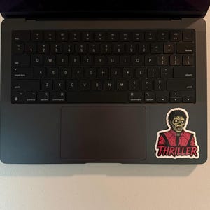 Może przedstawiać: Ciemnoszary laptop z czarną klawiaturą i gładzikiem. Na powierzchni laptopa naklejka z postacią zombie w czerwonej kurtce z napisem "THRILLER".