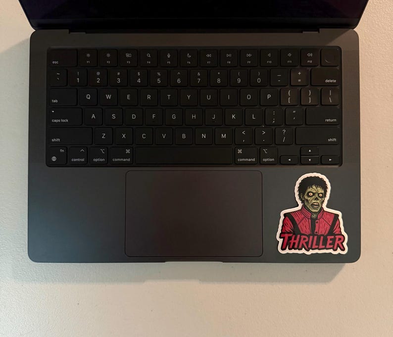 Może przedstawiać: Ciemnoszary laptop z czarną klawiaturą i gładzikiem. Na powierzchni laptopa naklejka z postacią zombie w czerwonej kurtce z napisem "THRILLER".