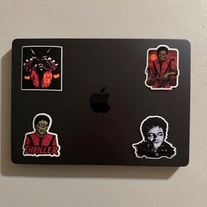 Może przedstawiać: Czarny laptop z czterema naklejkami przedstawiającymi Michaela Jacksona w jego postaci z Thrillera. Jedna naklejka ma napis "Thriller" w kolorze czerwonym. Inne naklejki przedstawiają potwora i czerwono-pomarańczowe tło.