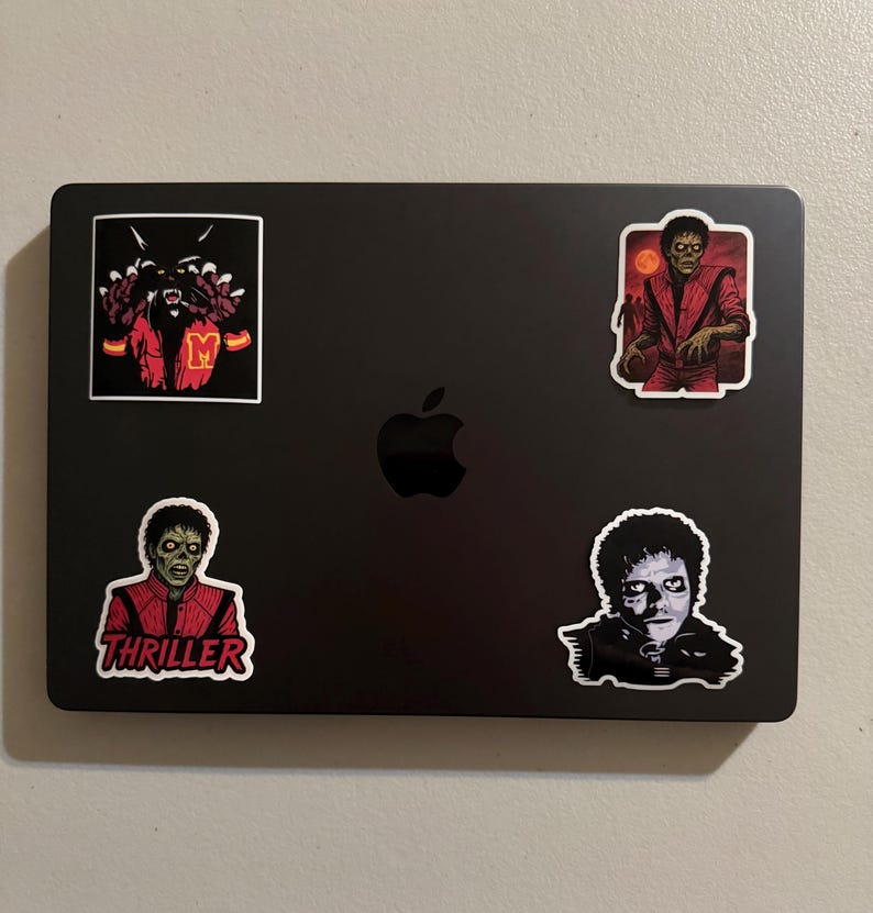 Może przedstawiać: Czarny laptop z czterema naklejkami przedstawiającymi Michaela Jacksona w jego postaci z Thrillera. Jedna naklejka ma napis "Thriller" w kolorze czerwonym. Inne naklejki przedstawiają potwora i czerwono-pomarańczowe tło.