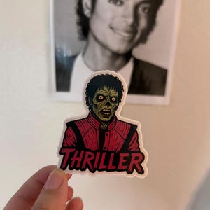 Może przedstawiać: Naklejka przedstawiająca Michaela Jacksona jako zombie w czerwonej kurtce. Słowo "THRILLER" jest napisane czerwonymi literami. W tle czarno-białe zdjęcie Michaela Jacksona.