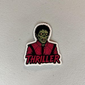 Może przedstawiać: Naklejka przedstawiająca postać podobną do zombie w czerwonej kurtce z napisem "Thriller" w czerwonym i białym tekście. Postać ma zieloną sk&oacute;rę, ż&oacute;łte oczy i czarne kręcone włosy. Naklejka jest na białym tle.