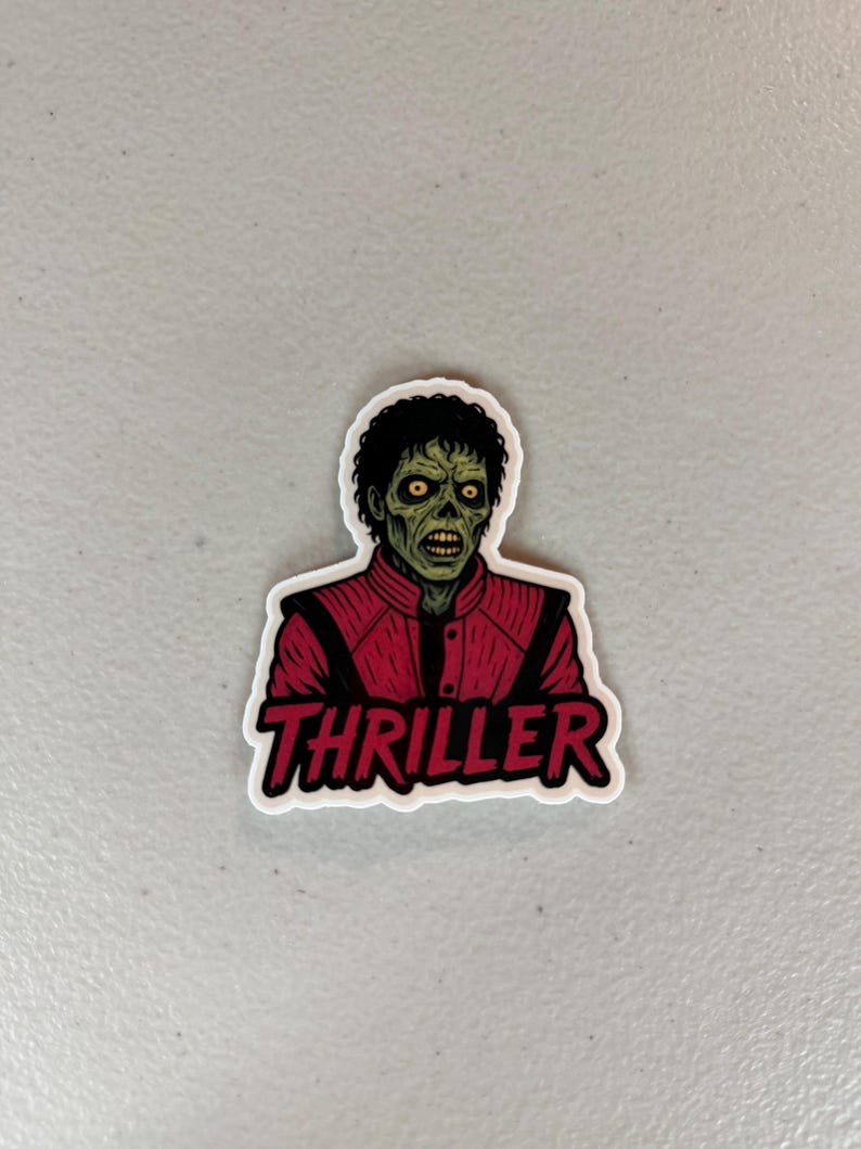 Może przedstawiać: Naklejka przedstawiająca postać podobną do zombie w czerwonej kurtce z napisem "Thriller" w czerwonym i białym tekście. Postać ma zieloną sk&oacute;rę, ż&oacute;łte oczy i czarne kręcone włosy. Naklejka jest na białym tle.