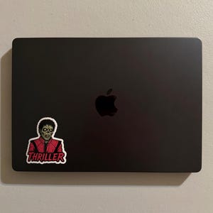 Może przedstawiać: Czarny laptop ze srebrnym logo Apple. Naklejka z Michaelem Jacksonem w kostiumie zombie z Thrillera jest przymocowana w lewym dolnym rogu. Naklejka zawiera słowo "Thriller" w czerwonym tekście.