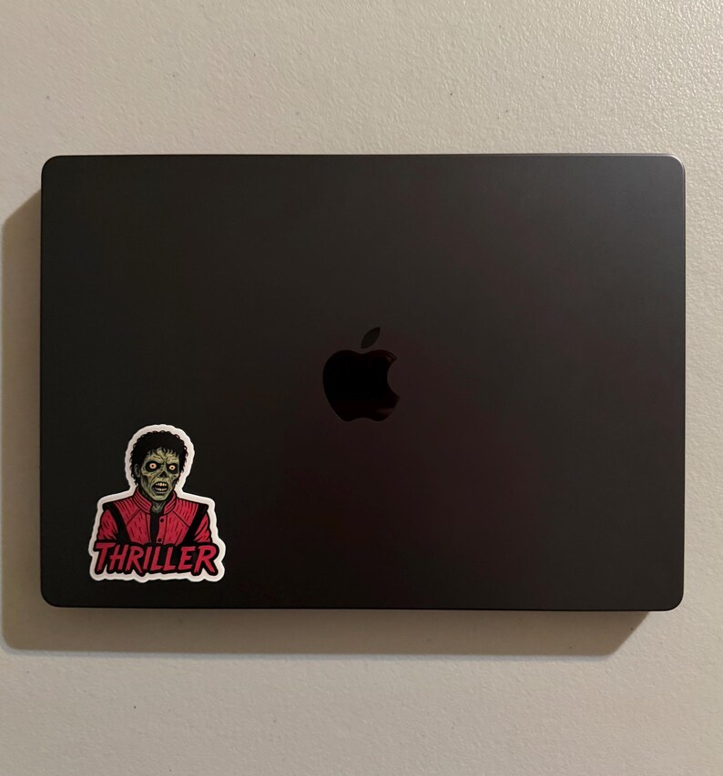 Może przedstawiać: Czarny laptop ze srebrnym logo Apple. Naklejka z Michaelem Jacksonem w kostiumie zombie z Thrillera jest przymocowana w lewym dolnym rogu. Naklejka zawiera słowo "Thriller" w czerwonym tekście.