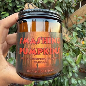 May include: An amber glass candle jar with a black screw-top lid. The label features a forest scene with the text SMASHING PUMPKINS, ORANGE, PUMPKIN, PACHOULI, VANILLA, and 8oz / up to 50 hr burn. The candle is held against a background of green leaves.