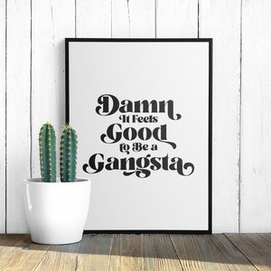 May include: Black and white typography print with the text "Damn It Feels Good To Be A Gangsta" in a retro font style. The print is framed and displayed on a white wood wall with a potted cactus in front of it.
