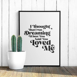 May include: Black and white typography print with the text "I thought that I was Dreaming When You Said You Loved Me". The print is framed in a black frame and is displayed on a white wood background with a potted cactus in front of it.