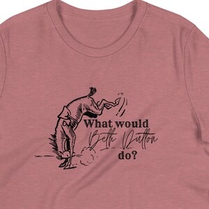 Könnte beinhalten: Ein staubrosa T-Shirt mit einer Schwarz-Weiß-Illustration einer Person auf einem wilden Pferd. Der Text "What would Beth Dutton do?" ist in einer Schriftart geschrieben. Das Shirt hat eine lockere Passform.