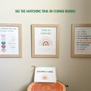 K&ouml;nnte beinhalten: Drei gerahmte Drucke mit einem Regenbogen-Design und Text &uuml;ber Gef&uuml;hle, Zeit-in-Ecke und positive Affirmationen. Ein Satz beruhigender Karten mit einem Regenbogen-Design liegt auf einer Holzoberfl&auml;che.