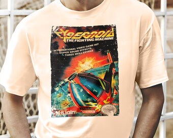 Puede incluir: Una camiseta de color melocotón claro con un diseño gráfico de videojuego vintage. El gráfico, titulado "Cyberoid: The Fighting Machine", muestra un vehículo futurista en rojo, azul y amarillo sobre un fondo espacial. El texto incluye "International Video Game Hit" y "Three Skill Levels".