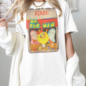Può includere: Maglietta bianca con una grafica vintage di cartuccia di gioco Atari 2600 con Mr. Pac-Man e fantasmi.