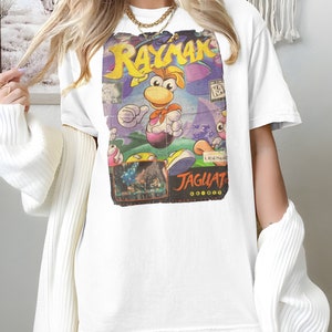 May include: White t-shirt with a vintage Rayman video game graphic printed on the front. The graphic features a cartoon character with a purple and yellow color scheme. The text "Rayman" and "Jaguar" are visible on the graphic.