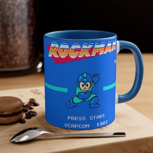 May include: A blue ceramic mug featuring a blue and white image of the video game character Rockman. The mug displays the text "ROCKMAN" in a retro video game style font. The image also includes the text "PRESS START", "@CAPCOM", and "1987".