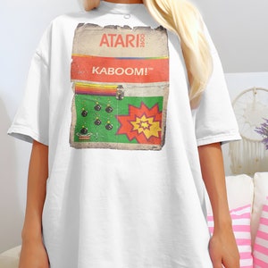 Puede incluir: Camiseta blanca de gran tamaño con un diseño de cartucho de juego Atari 2600 vintage. El diseño presenta el texto "ATARI 2600" y "KABOOM!" con un gráfico verde y rojo de un videojuego.