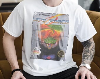 Puede incluir: Camiseta blanca con un gráfico retro del videojuego "Life Force". El diseño muestra el título del juego, una colorida ilustración de una criatura y el logotipo de Nintendo. La camiseta está hecha de un material suave.