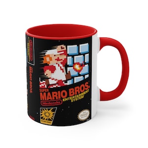 Puede incluir: Taza de cerámica blanca con interior y asa rojos. La taza presenta un diseño de videojuego retro con fondo negro, texto blanco y rojo y una imagen pixelada de Mario del videojuego Super Mario Bros. El texto en la taza dice "Super Mario Bros. Entertainment System Nintendo".