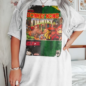 May include: White t-shirt with a vintage Donkey Kong Country video game cartridge graphic. The graphic is in color and features the characters Donkey Kong, Diddy Kong, and King K. Rool.