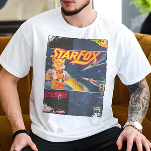 Pode incluir: Camiseta branca com um gráfico vintage de um cartucho de videogame Star Fox. O gráfico apresenta um personagem de raposa, naves espaciais e o texto "Star Fox" e "Super Nintendo".