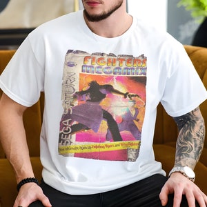 May include: White t-shirt featuring a vintage-style graphic for the video game Fighters Megamix. The design includes the Sega Saturn logo and the text "the ultimate team-up-Fighting Vipers and Virtua Fighters". The graphic has a distressed, aged appearance.
