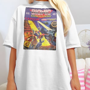 May include: White t-shirt with a graphic print of a vintage video game cartridge. The game is called "Bionic Commando" and is made by Capcom. The image shows a character in a red and blue suit fighting a robot.
